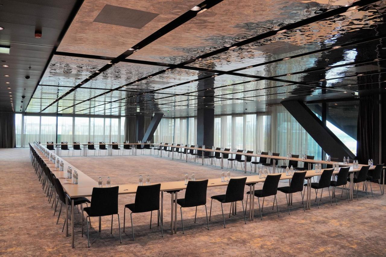 Melia Vienna Hotel Exterior photo The photo shows a modern conference room with a long rectangular table arranged in a U-shape. The table is flanked by multiple black chairs. The room features a sleek, reflective ceiling that gives a contemporary feel, and large windows allow natural