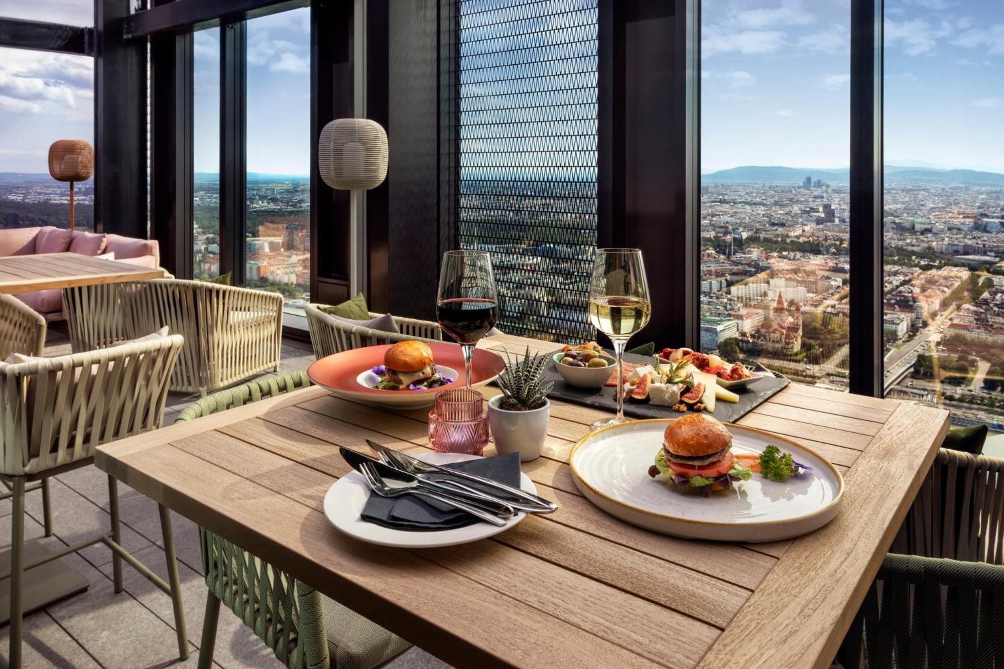 Melia Vienna Hotel Exterior photo The photo shows a stylish dining setup on a high rooftop or terrace, with a panoramic view of a city skyline in the background. 

There are two tables made of wood; one features two glasses of white wine and an assortment of dishes, including a plate