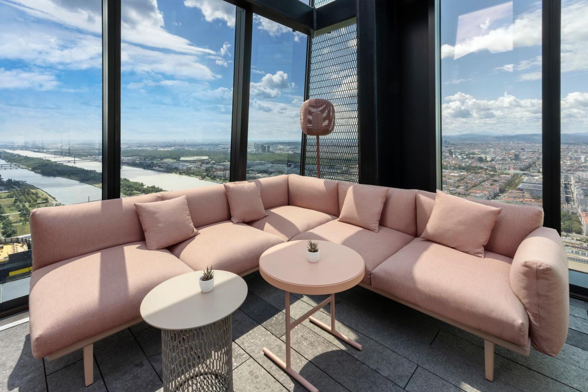 Melia Vienna Hotel Exterior photo The photo shows a modern lounge area with a large, L-shaped pink couch adorned with several soft cushions. In front of the couch, there are two tables: one round and one with a more intricate design. The space is located in a high-rise building, offe