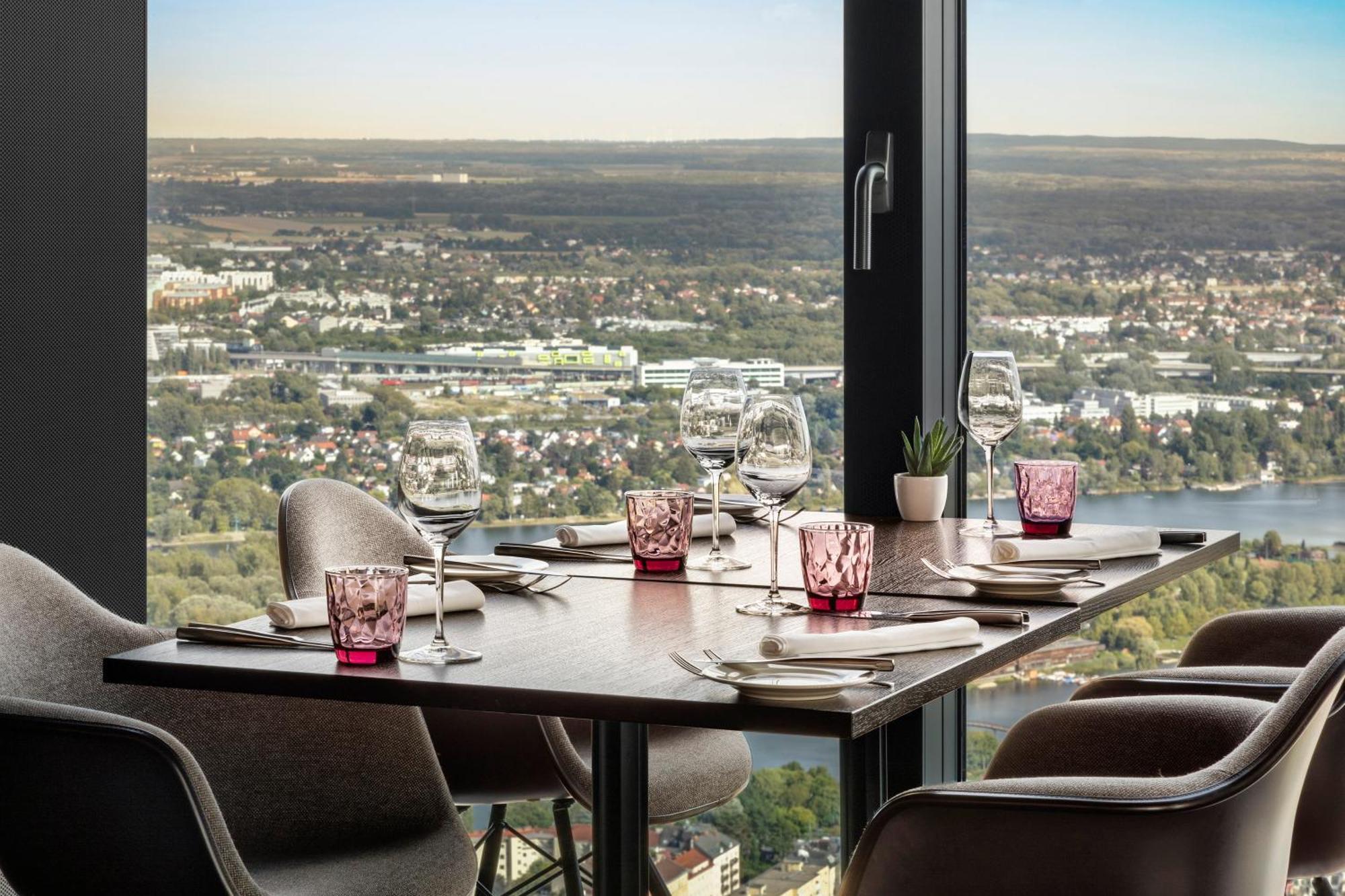 Melia Vienna Hotel Exterior photo The photo shows a dining table set up in a restaurant with a view. The table has dark wood and features several dining essentials arranged neatly. There are elegant wine glasses, pink-tinted glass holders, and plates set beside them. A small plant is