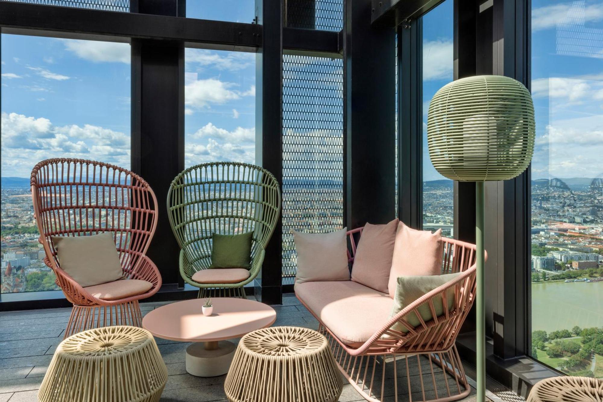 Melia Vienna Hotel Exterior photo The photo shows a modern lounge area with large windows offering a view of the outdoors. There are two stylish, high-backed chairs made of wicker in pastel colors—one pink and one green. A cozy sofa in a soft pink hue is positioned between them, acco
