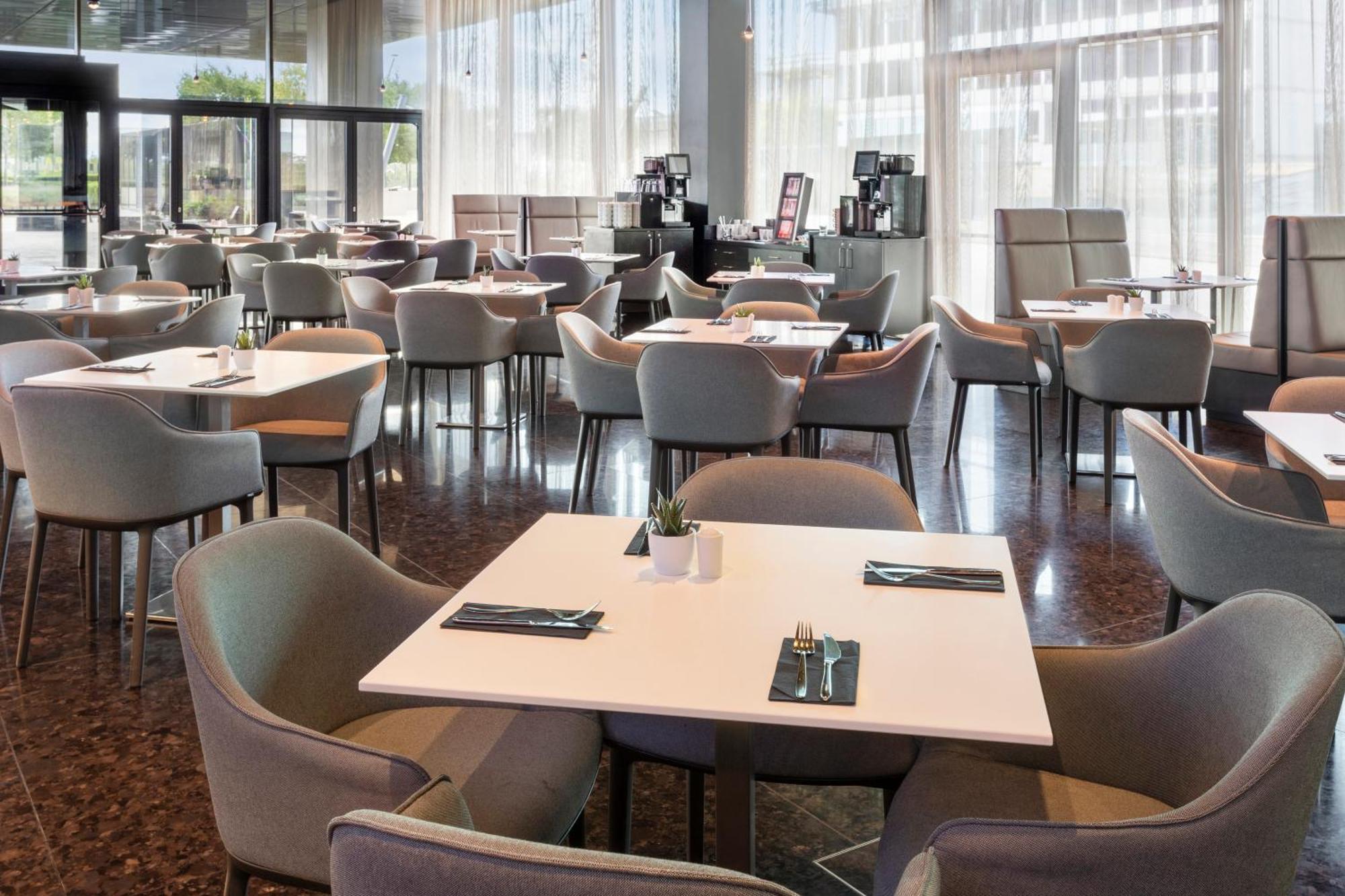 Melia Vienna Hotel Exterior photo The photo shows a spacious dining area in a restaurant or café. There are several tables arranged throughout the space, each with light-colored tabletops and comfortable chairs. The decor appears modern and minimalistic, with large windows allowing n
